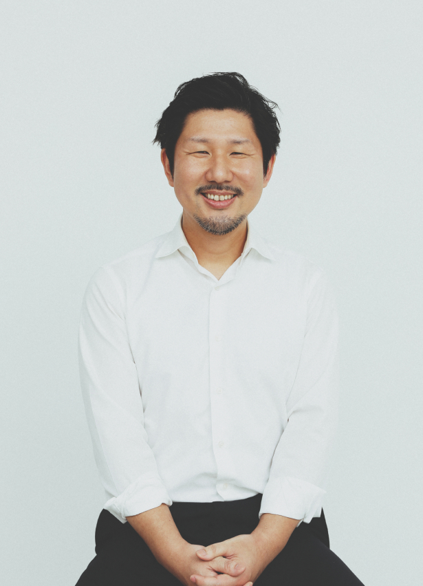 大硲 神。白いシャツで椅子に腰かけ、こちらを見て笑っている